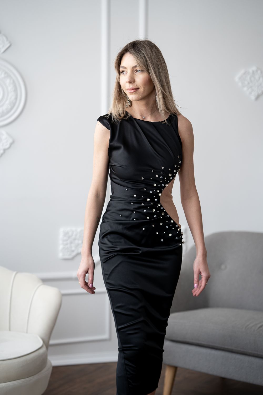 Model showcases black dress with pearl embellishments in minimalist white room setting.