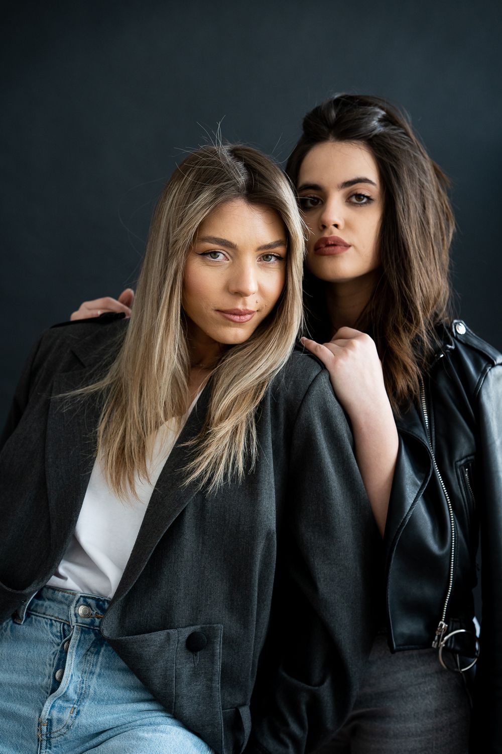 Two models pose in casual black outfits against dark background during fashion photoshoot.