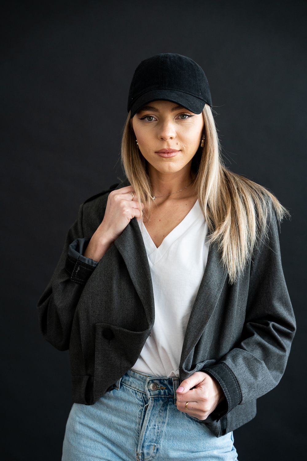 Casual fashion model wearing baseball cap, white shirt and dark blazer poses against black background.