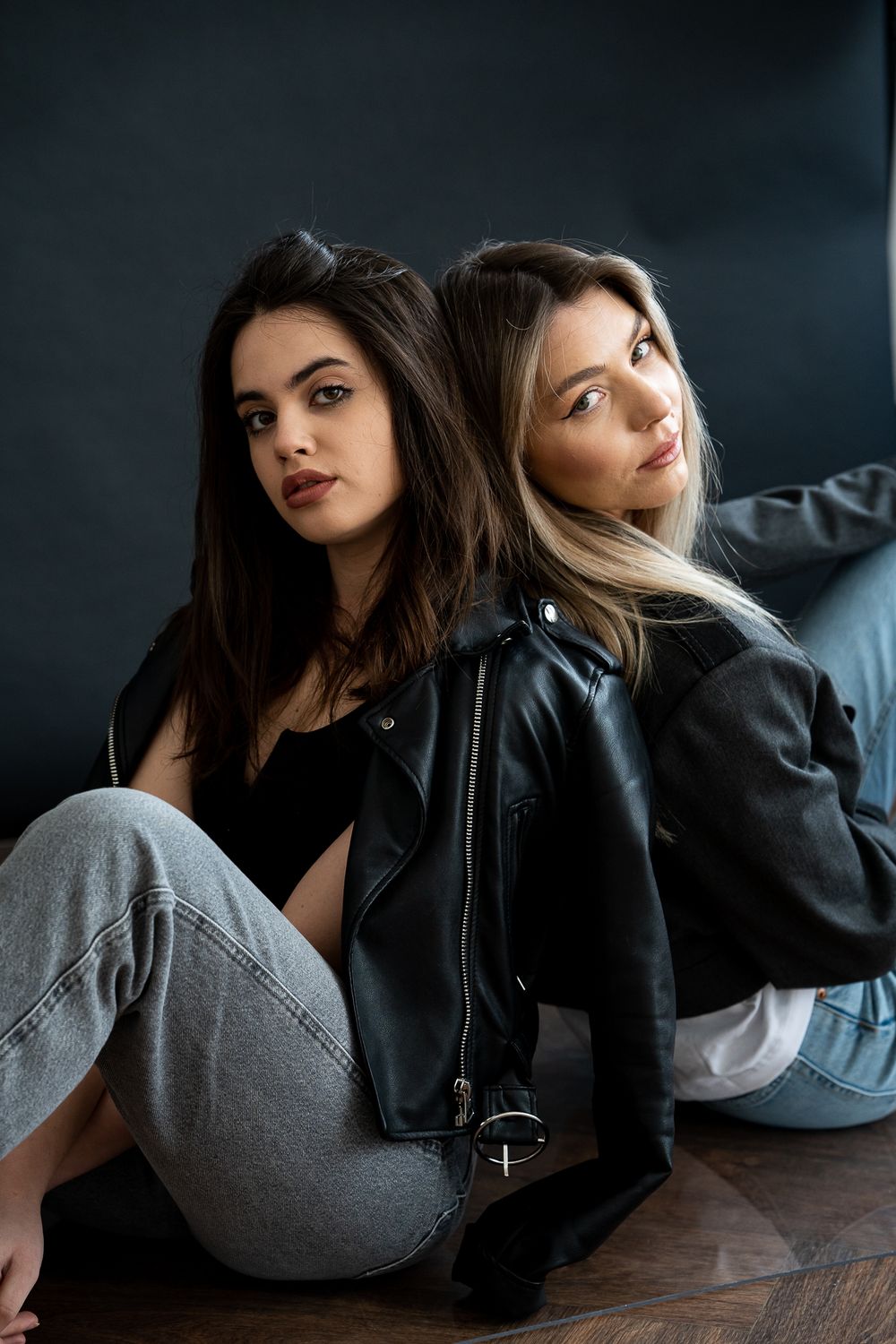 Two women in leather jackets and jeans pose against dark background.