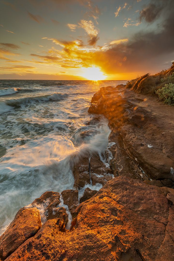 Dramatic coastal sunset with waves crashing against orange rocky cliffs under golden sky.