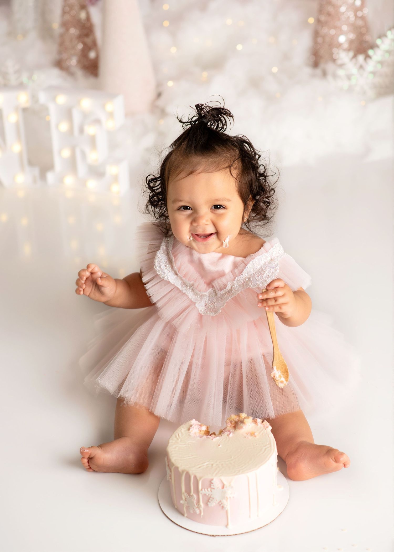 a baby smiles with a spoon covered in frosting during her smash cake winter onederland photoshoot