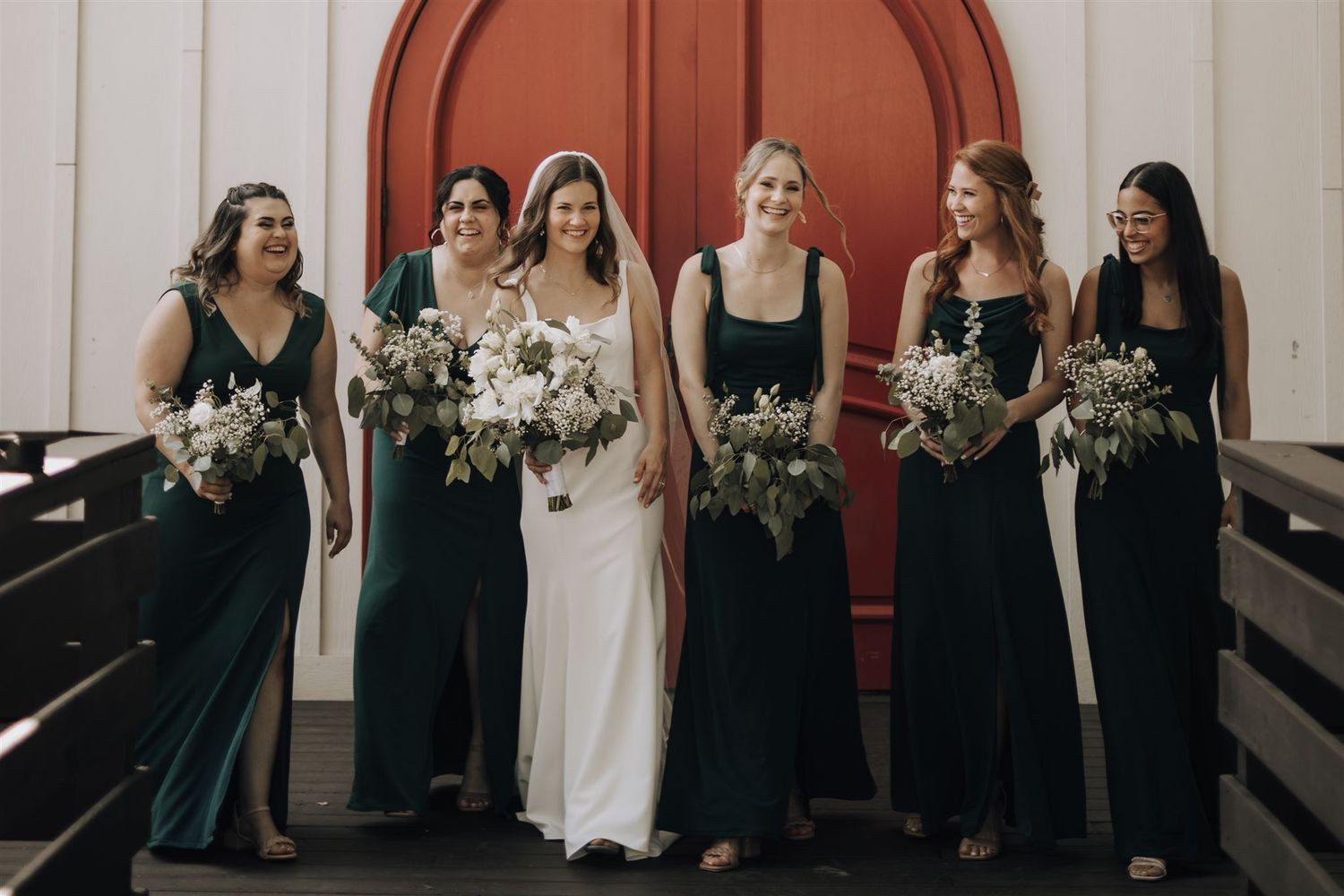 Wedding party stands together on church steps, featuring emerald gowns and white floral arrangements.