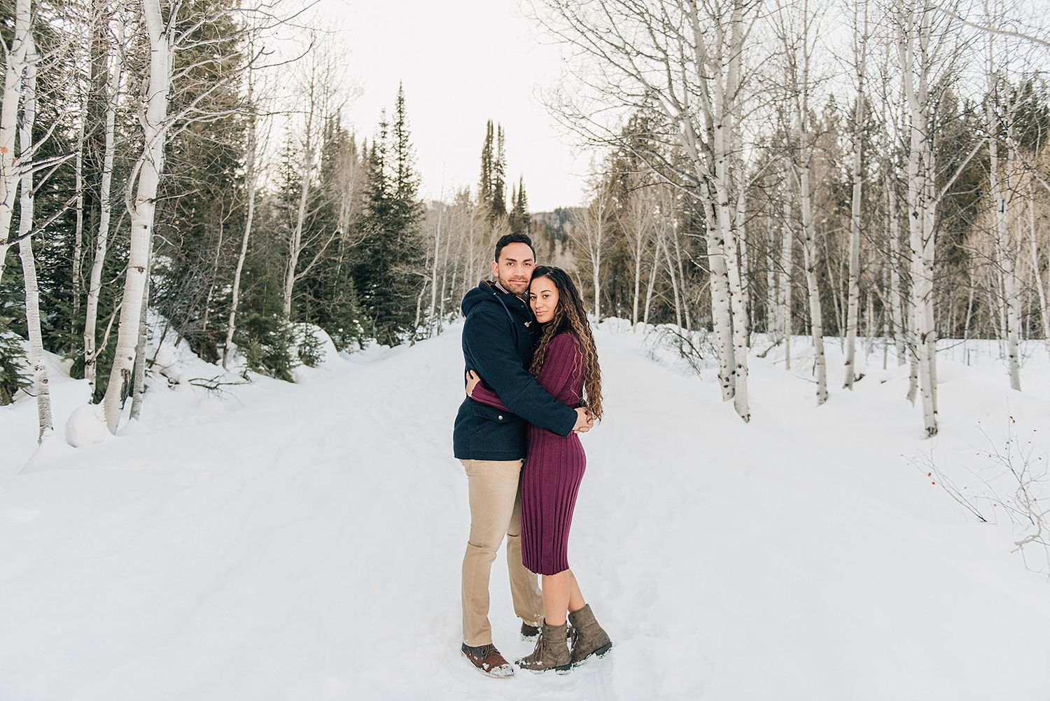 A couple embraces while standing in a snow-covered forest path during winter.