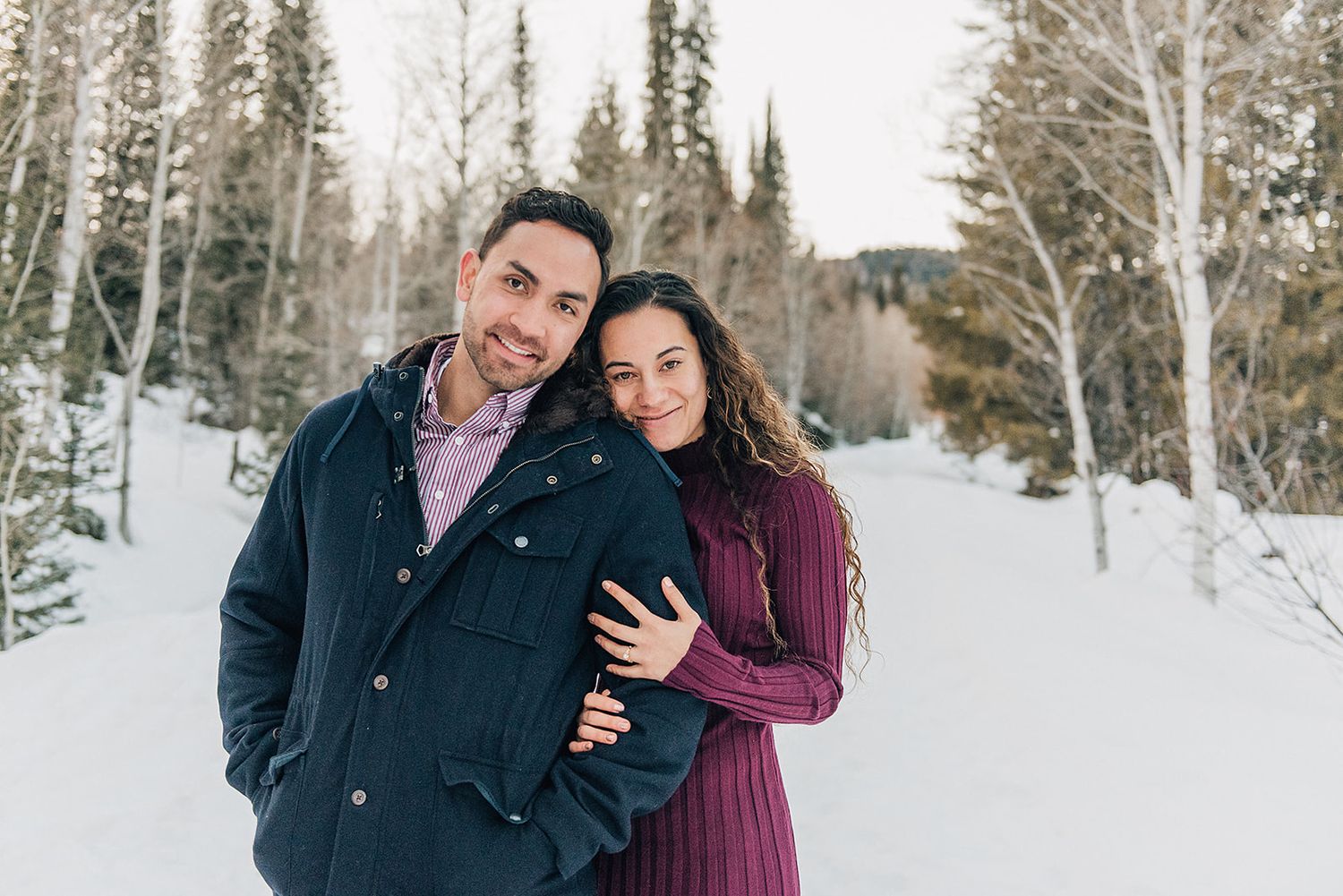 A couple poses in a snowy winter forest scene wearing coordinating burgundy and navy outfits.