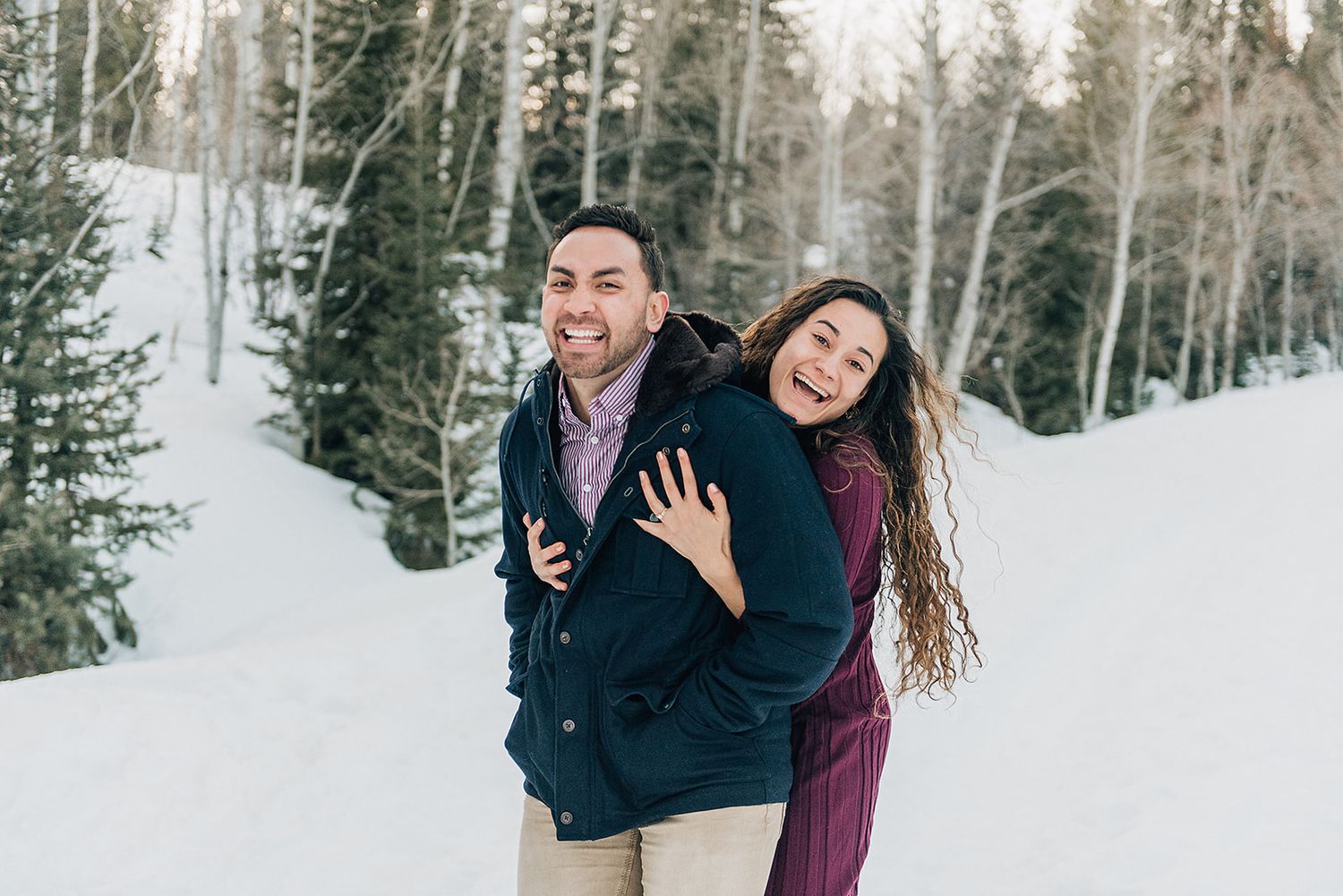 Playful winter engagement photo in snowy forest with pine trees.