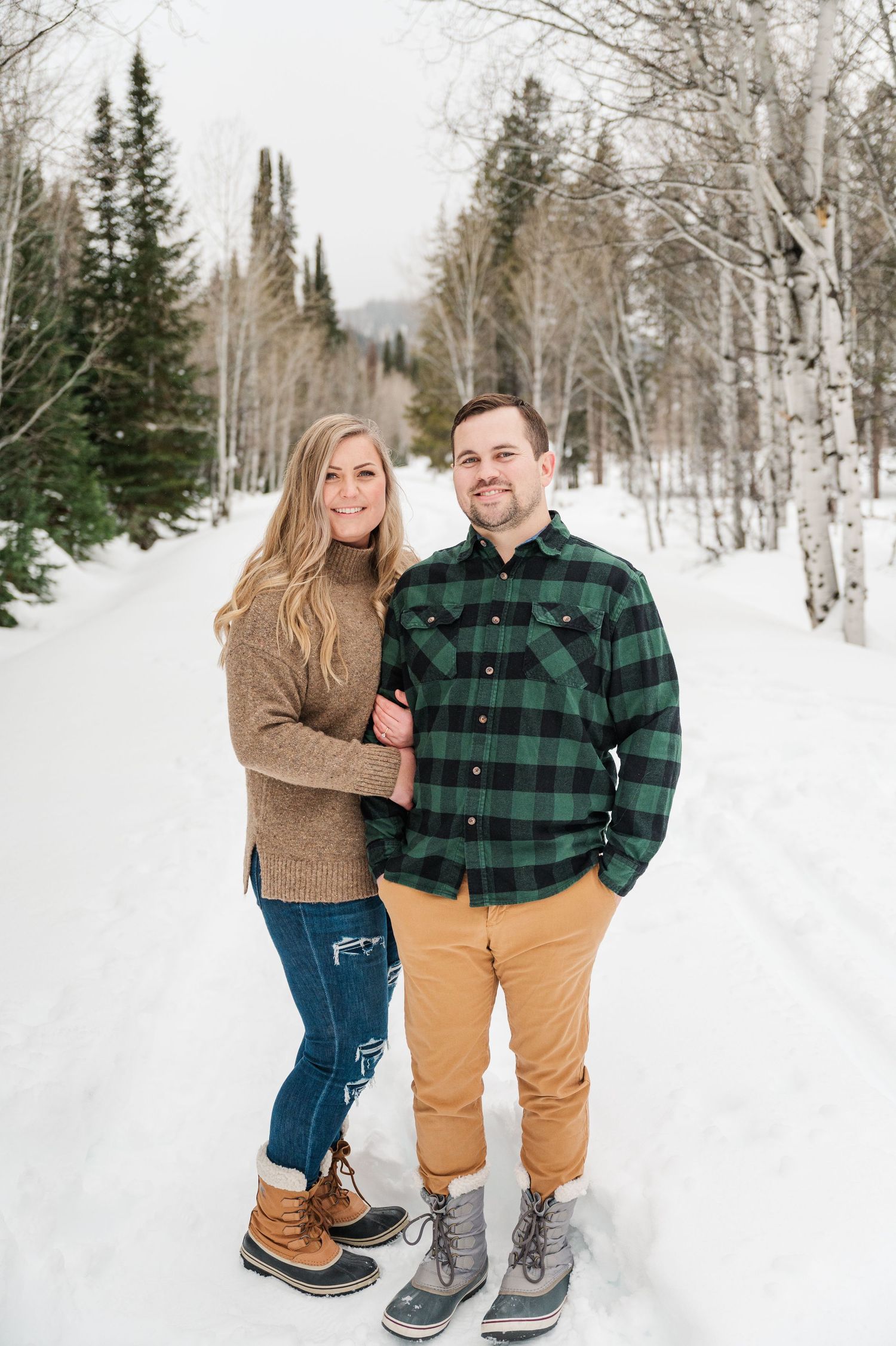 A couple in casual winter attire standing on a snow-covered path lined with birch trees.