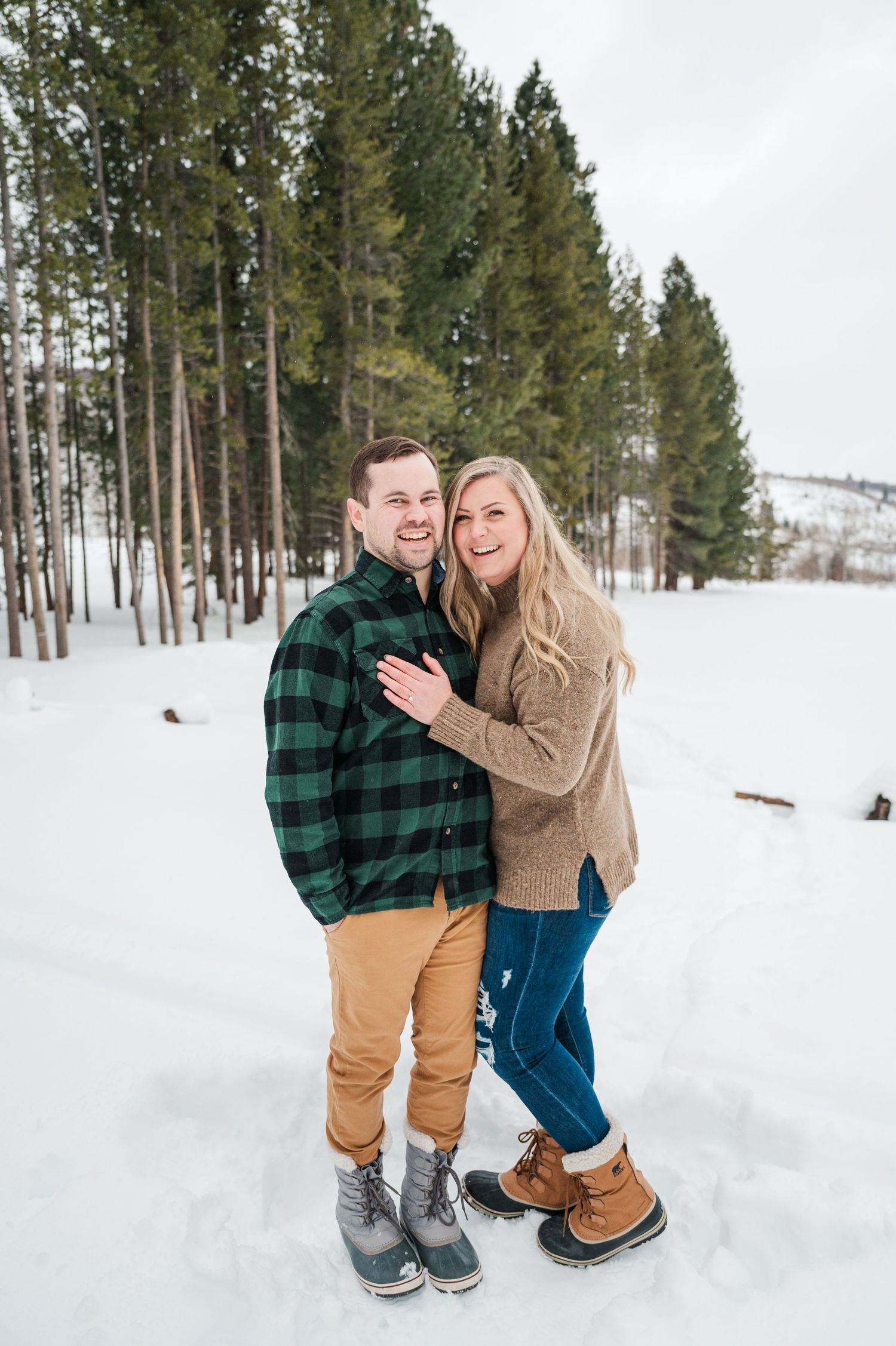 A couple in winter clothing posing in a snow-covered forest landscape.