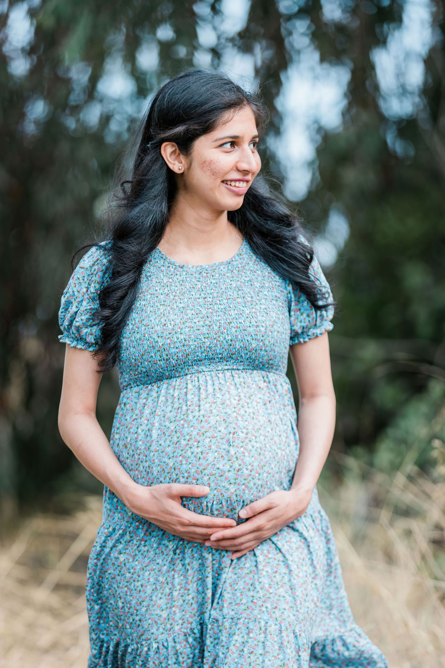 Expectant mother in a blue patterned dress smiling outdoors.