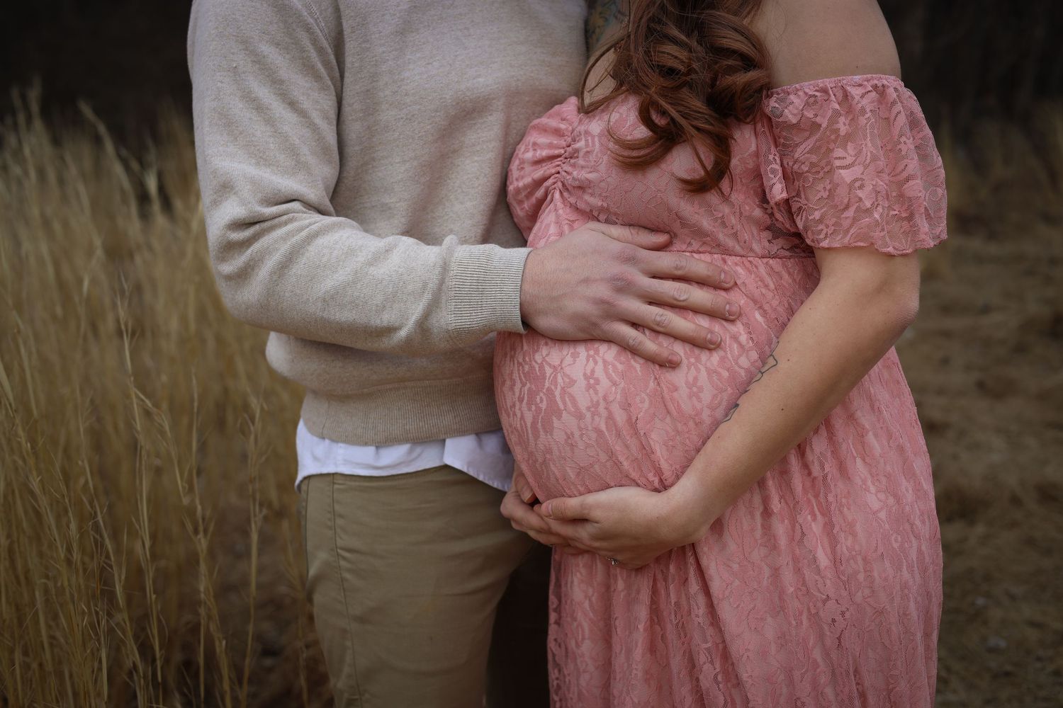 Maternity photo session in a field at sunset showing a couple embracing in soft pink and neutral tones.