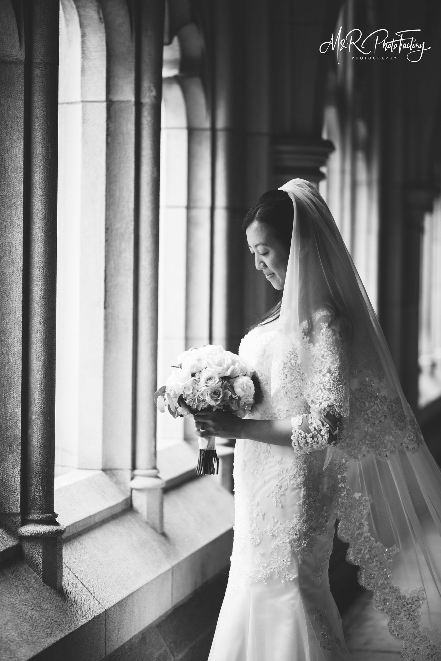 Elegant bride in lace gown holds a bouquet while standing by arched windows in black and white.