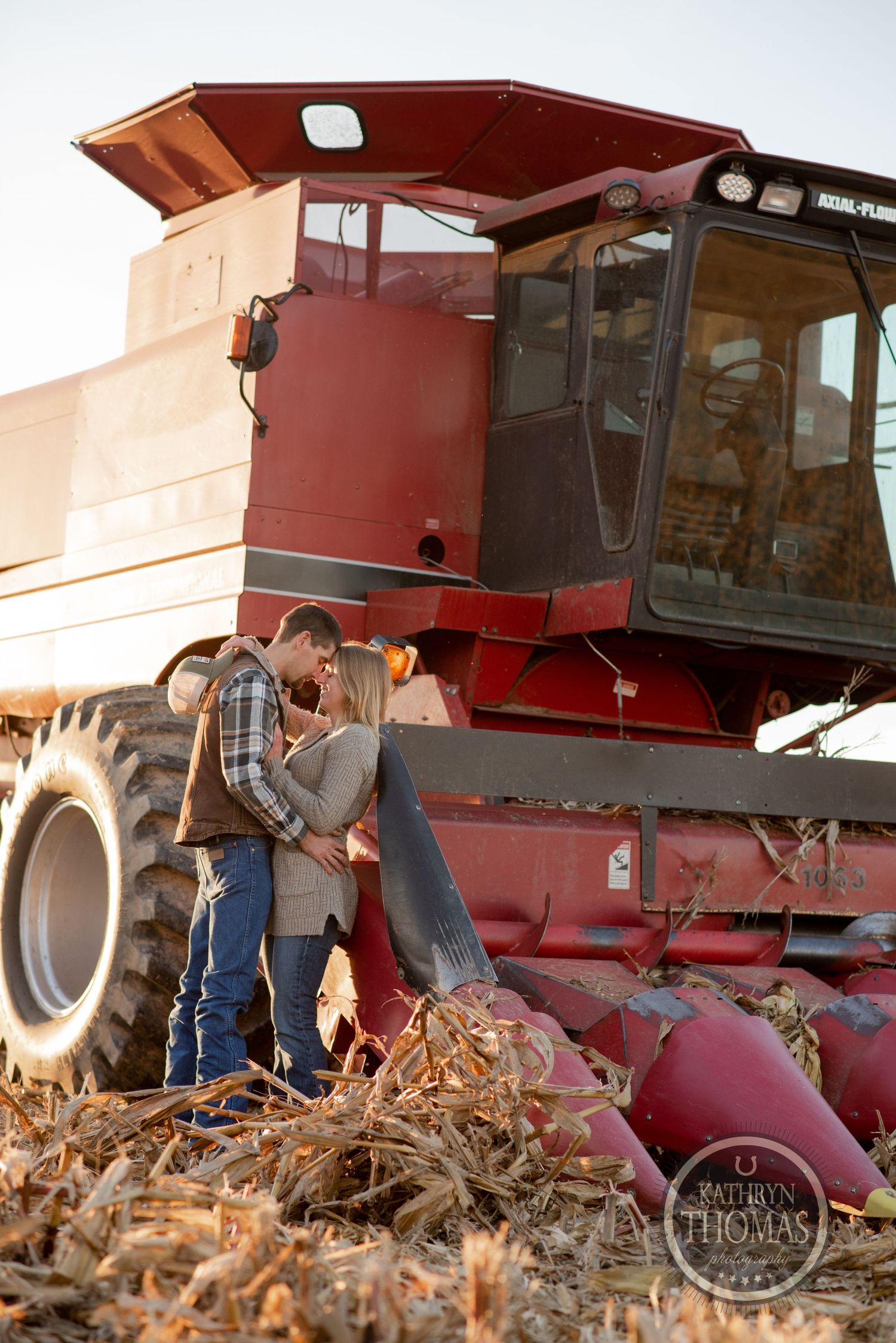 Romantic couple sharing a moment beside a red combine harvester in a harvested field.
