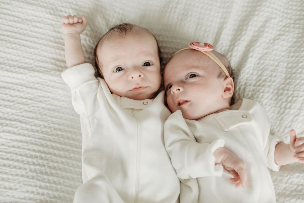Twin infants wearing white outfits laying side by side on white bedding.