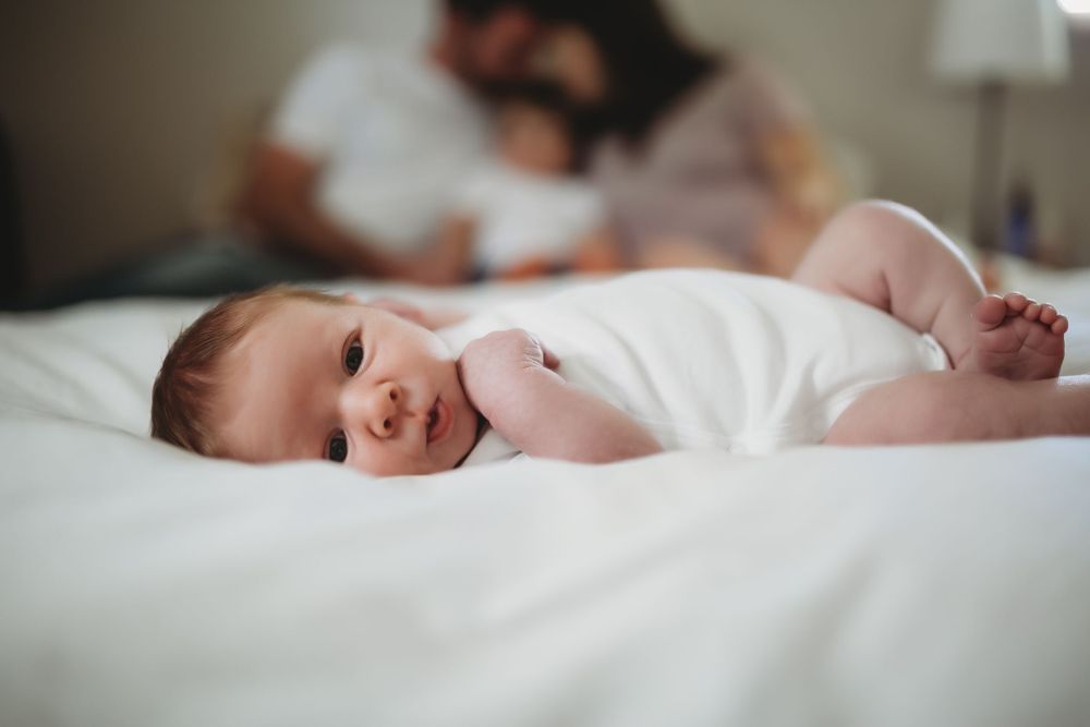A happy baby lies on white bedding while the family sits in soft focus in the background.