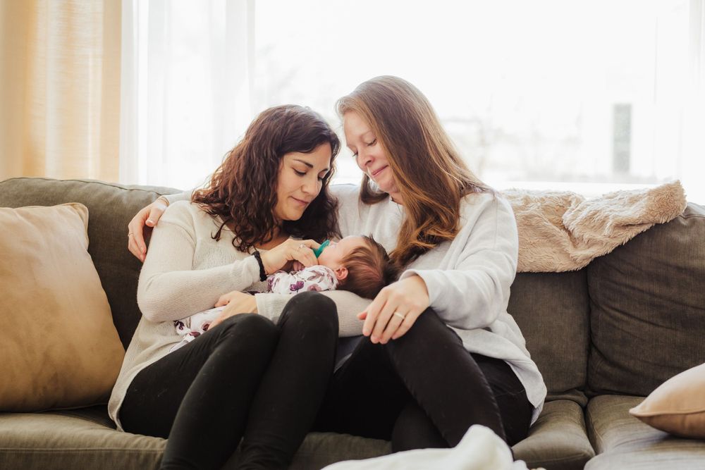 Two moms in cozy white sweaters share a relaxed moment together holding their new baby on a brown sofa with pillows.