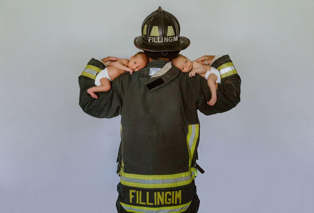 A firefighter in protective gear holds his twin babies while wearing a dark helmet with reflective strips.