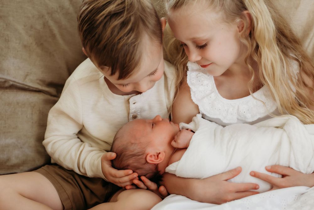 Three siblings sharing a tender moment on a beige couch with the older ones leaning over their baby sibling.