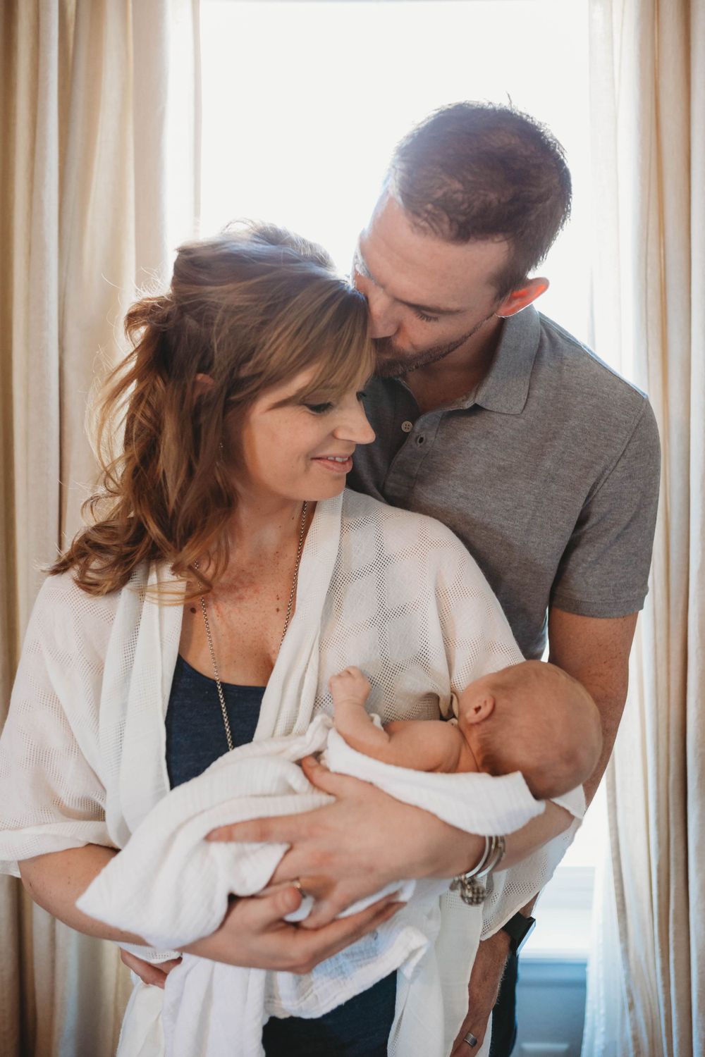 A tender family moment as parents hold their newborn baby wrapped in white while standing by a window with curtains.