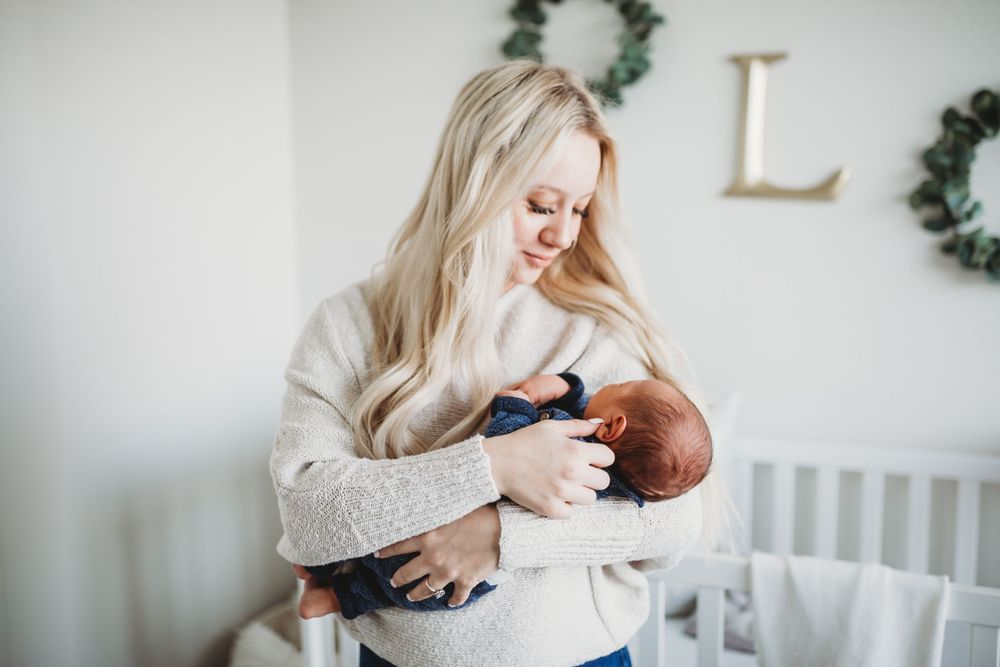 A mom in a cream sweater holds a newborn baby in a nursery decorated with white walls and decorative wreaths.