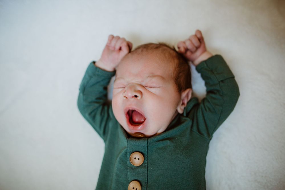 A yawning baby stretches while wearing a green button-up onesie against a white background.