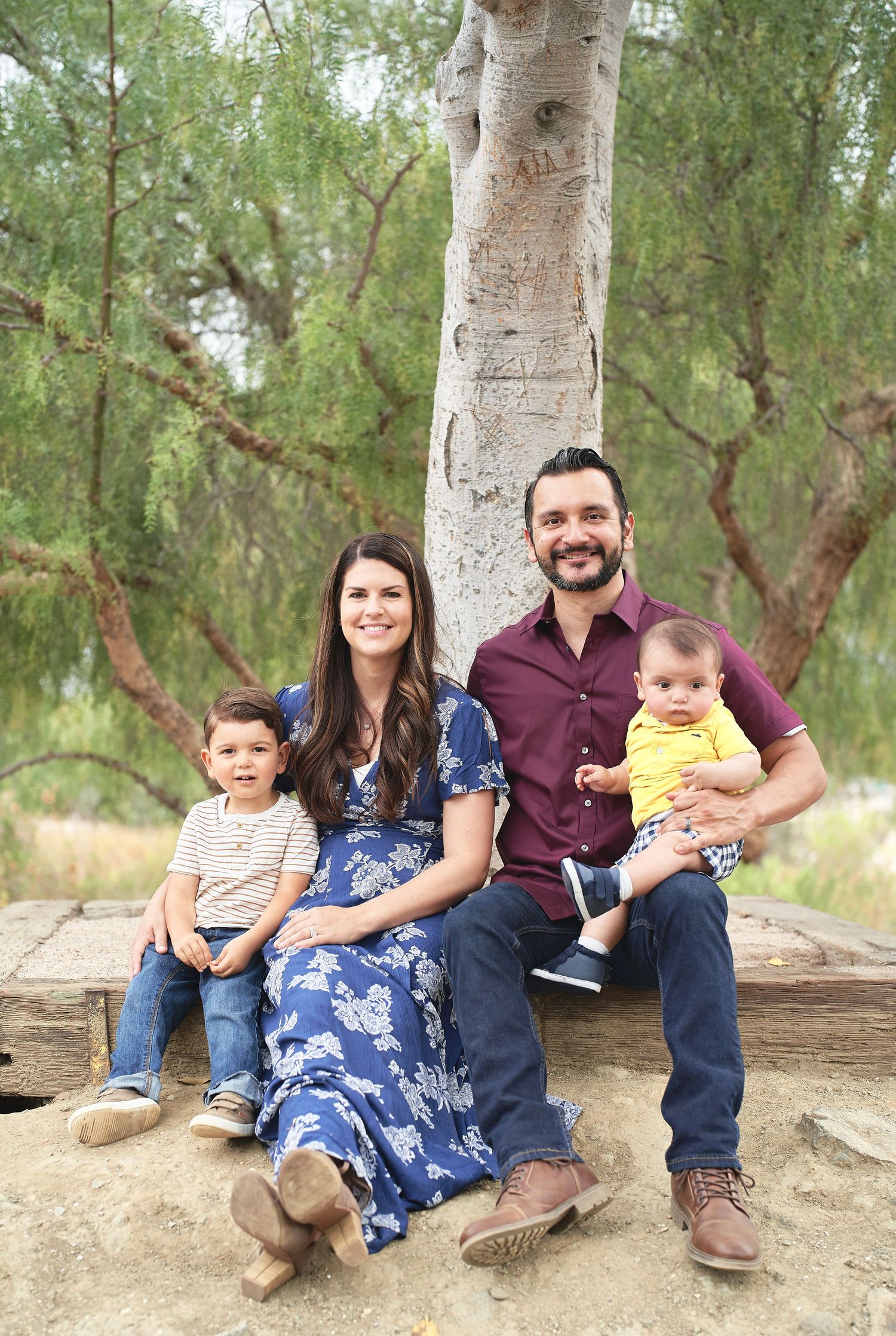 Family sits together on wooden bench under large birch tree.