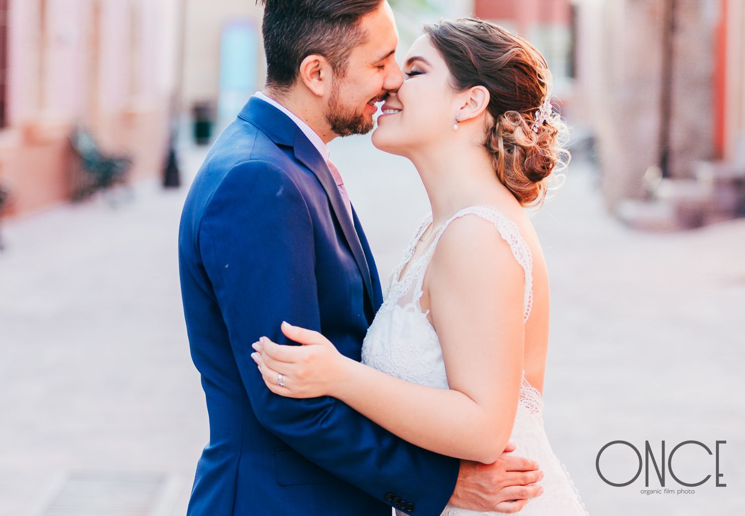 A tender moment between a couple in formal attire on a pink-hued street.