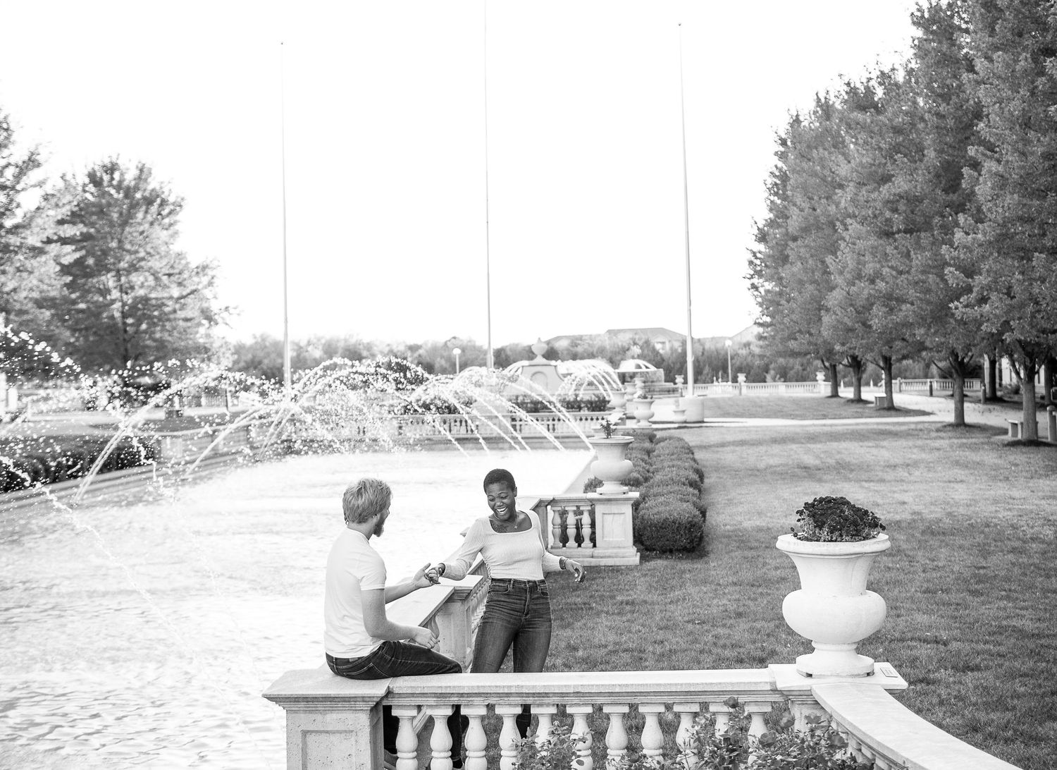 Two people sit on stone balustrade overlooking scenic landscape with trees in black and white photograph.