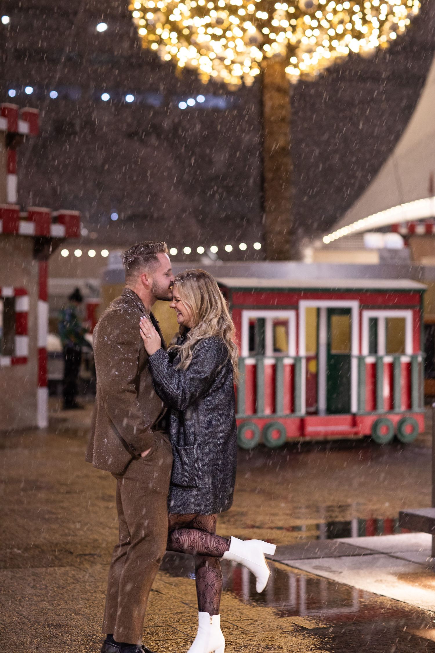 Couple sharing romantic moment in falling snow with festive lights and carnival backdrop.