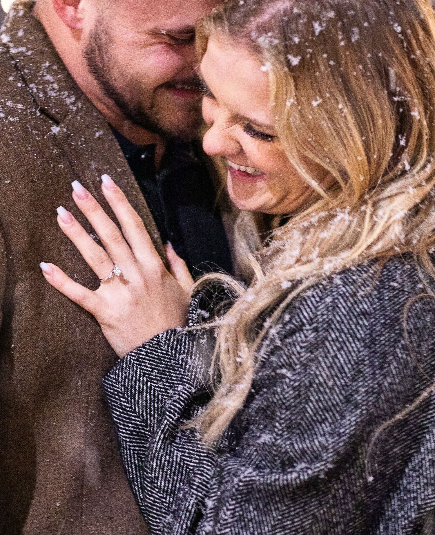 Close up of couple showing engagement ring while snow falls around them.