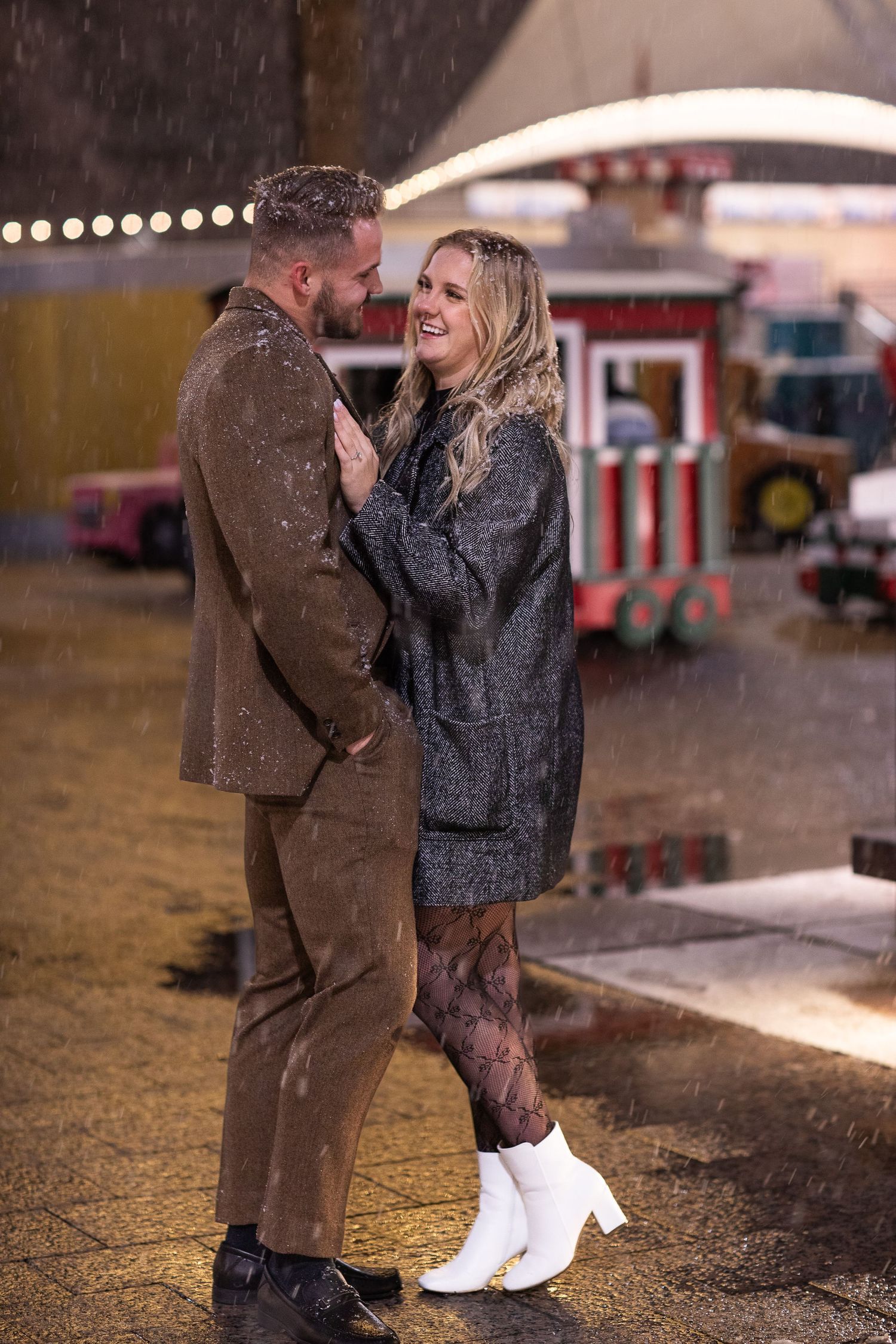 Happy couple laughing together in snowfall with carnival decorations behind them.