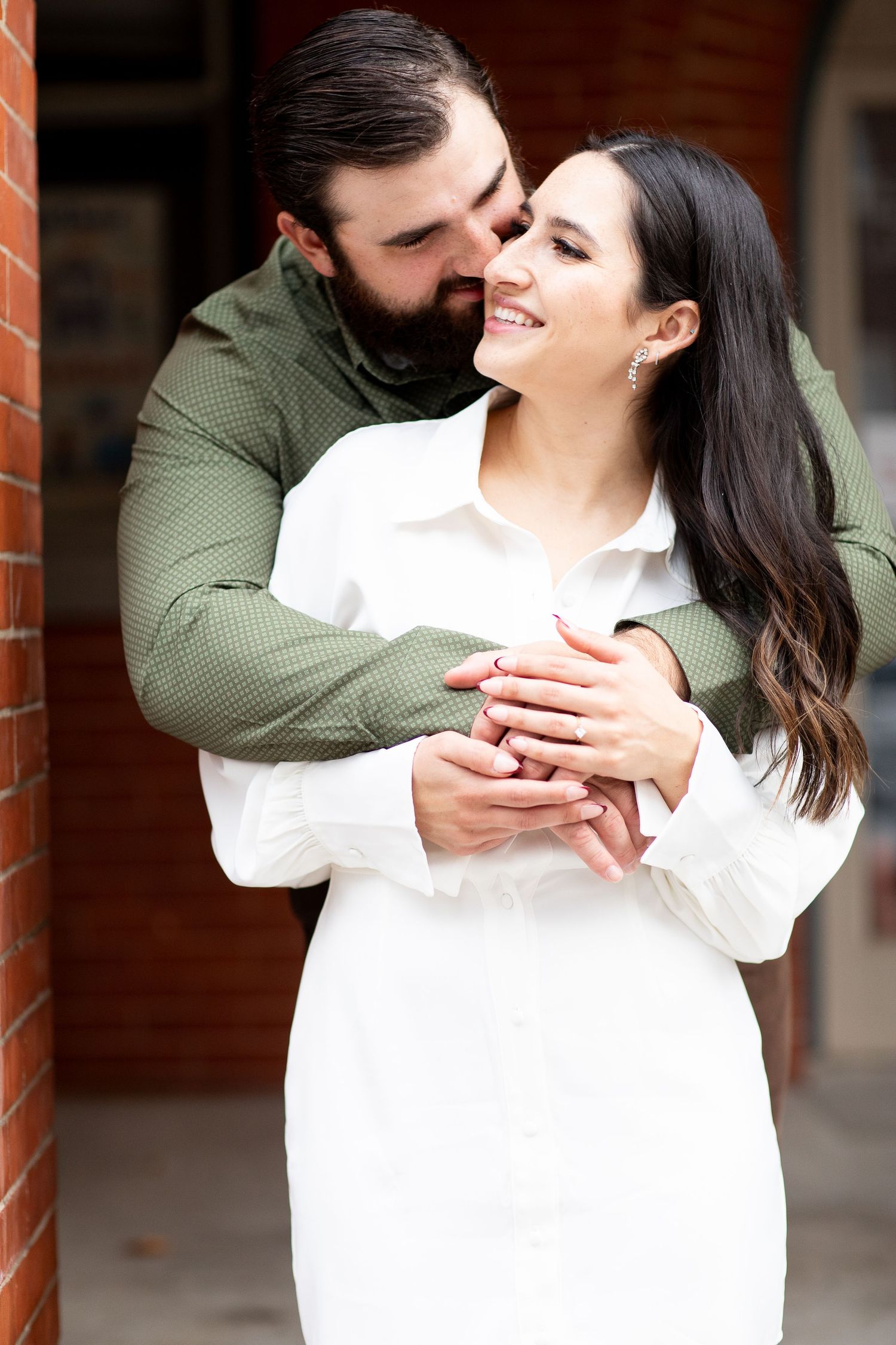 A romantic embrace between a couple where she wears white and he wears olive green against a brick wall background.