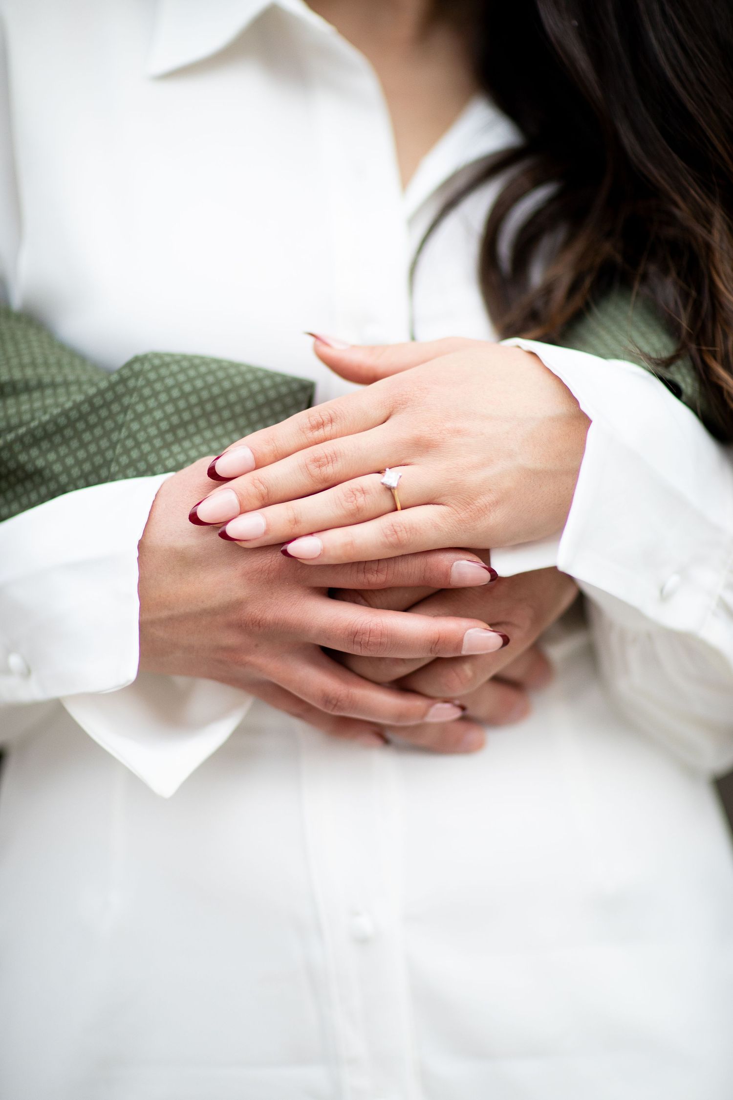 Close up of hands clasped together showing an engagement ring against white and olive green clothing.