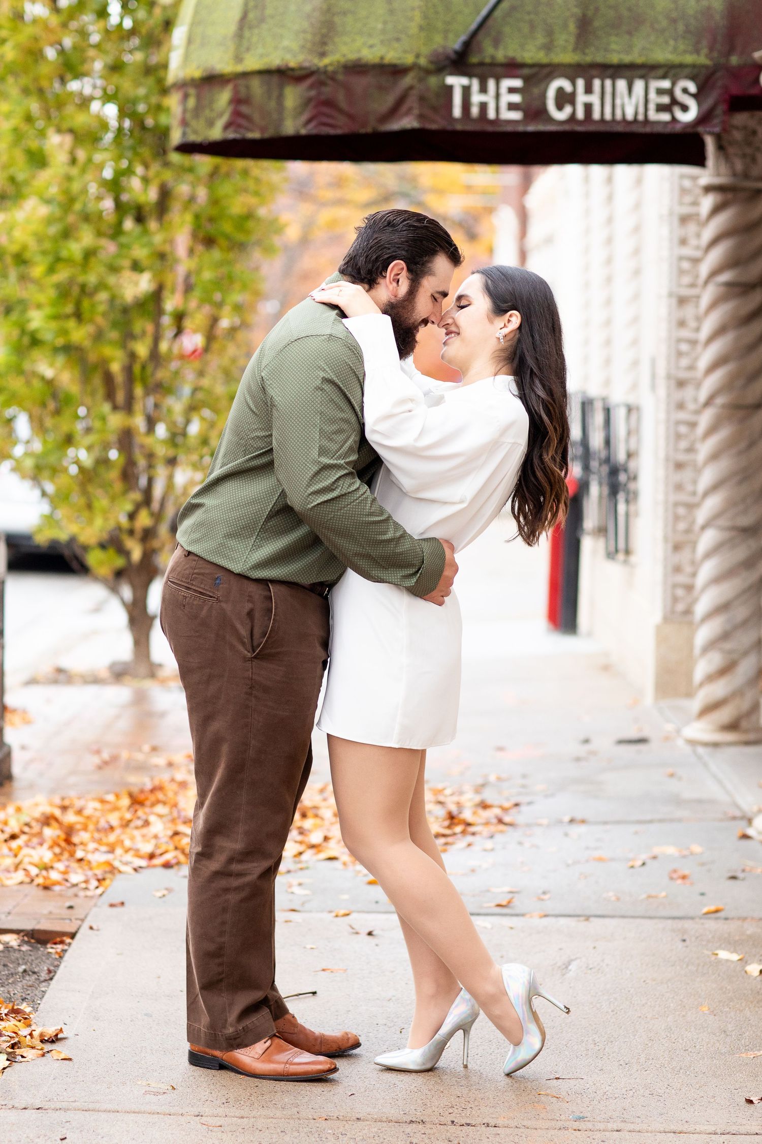 Couple in autumn sharing a romantic moment under The Chimes sign on a leaf-covered sidewalk.