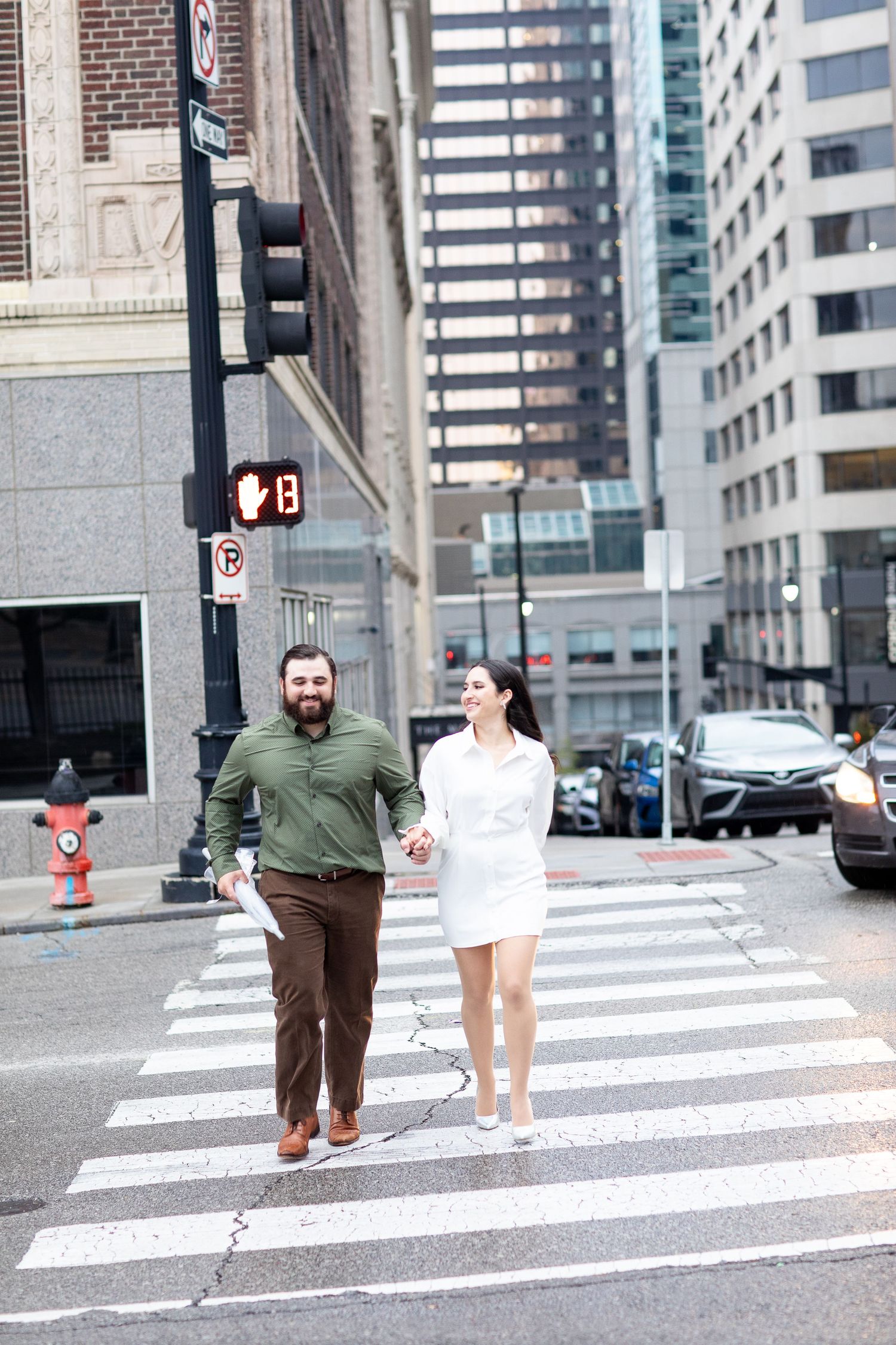 Couple walking hand in hand across an urban crosswalk with city buildings in the background.