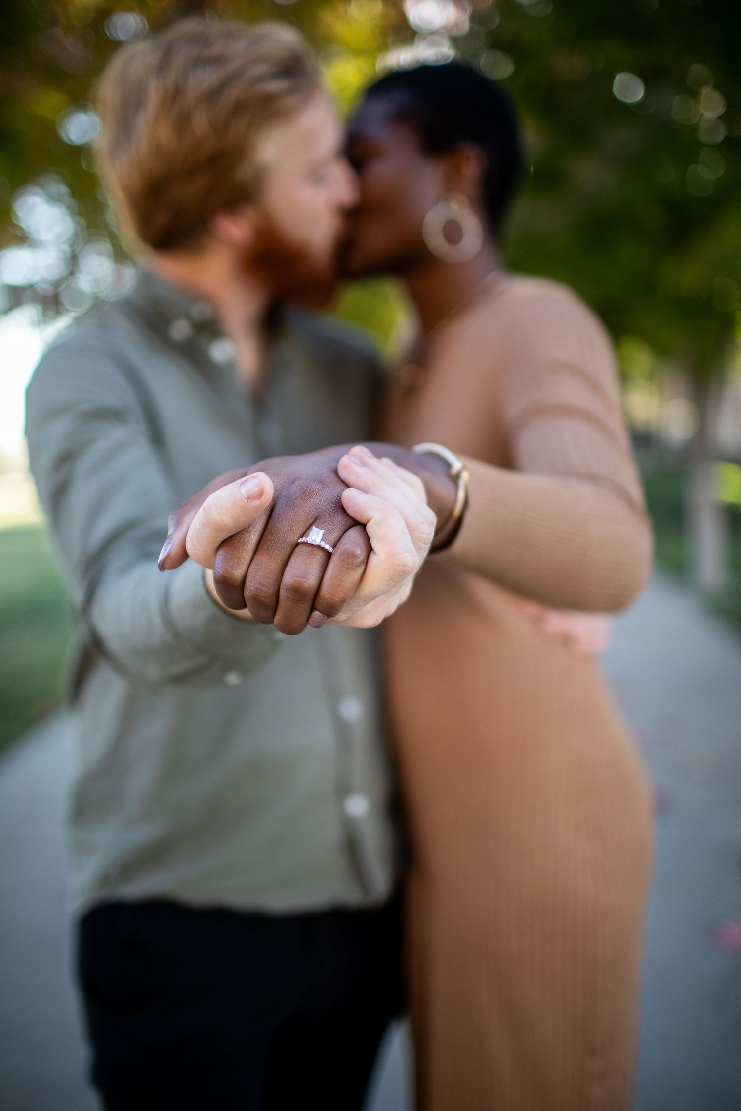 Close up of two hands holding each other with focus on engagement rings in an outdoor setting.