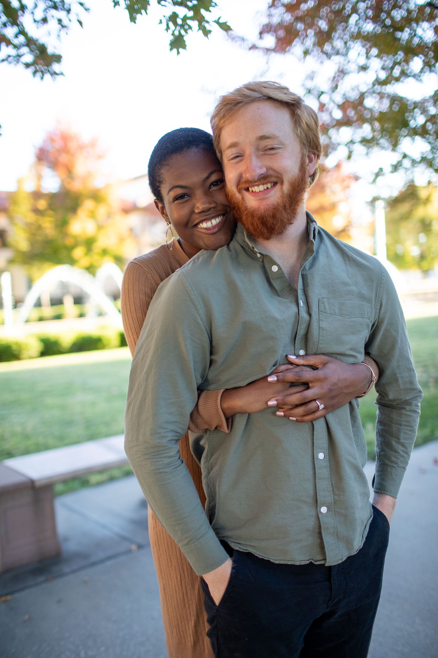Happy couple embracing outdoors with autumn trees and white archways in the background.