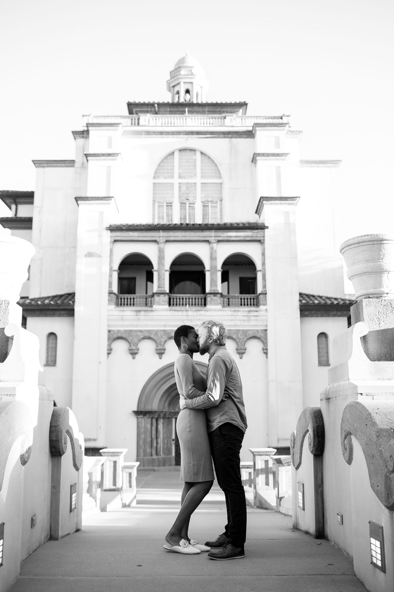 Black and white photo of couple embracing on bridge in front of Spanish-style building architecture.