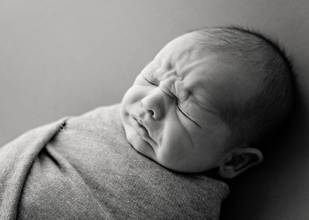 Black and white close-up of newborn making expressive face while swaddled in gray wrap.
