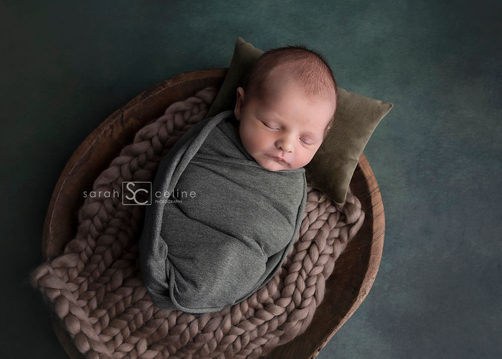 Sleeping newborn wrapped in gray swaddle resting in wooden bowl with knit blanket.