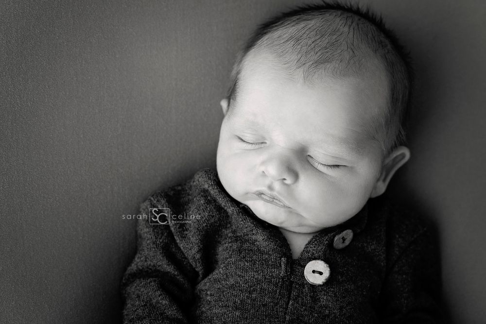 Black and white portrait of sleeping newborn in buttoned sweater against dark backdrop.