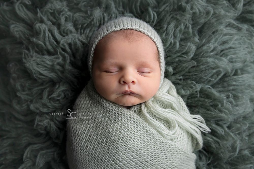 Sleeping newborn wrapped in gray knit blanket wearing matching hat on furry backdrop.