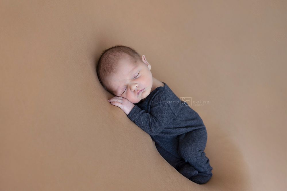 Newborn baby in dark outfit sleeping peacefully on tan colored background.