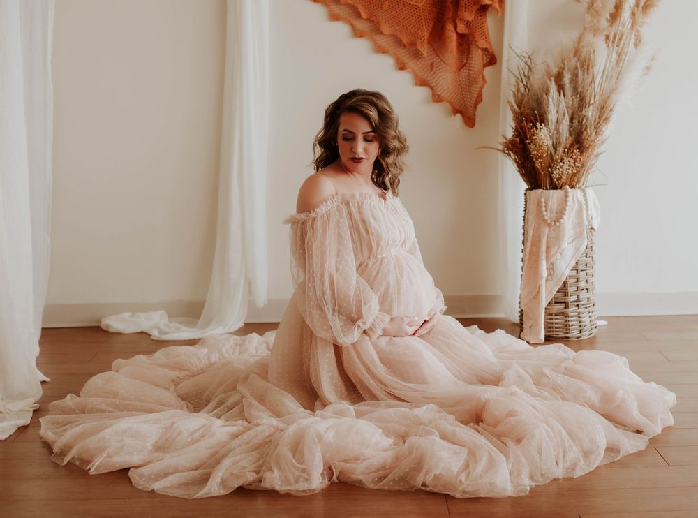 A pregnant woman in a flowing blush ruffled tulle gown sits elegantly on a studio floor.