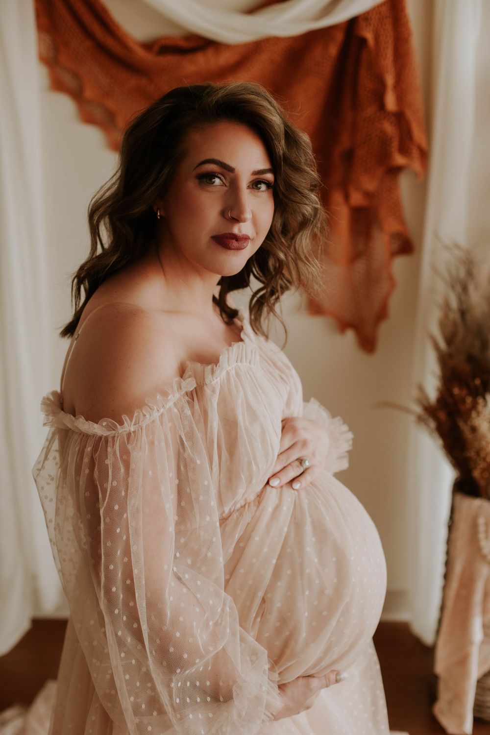 A pregnant woman in an off-shoulder blush tulle maternity gown poses gracefully in a boho studio.