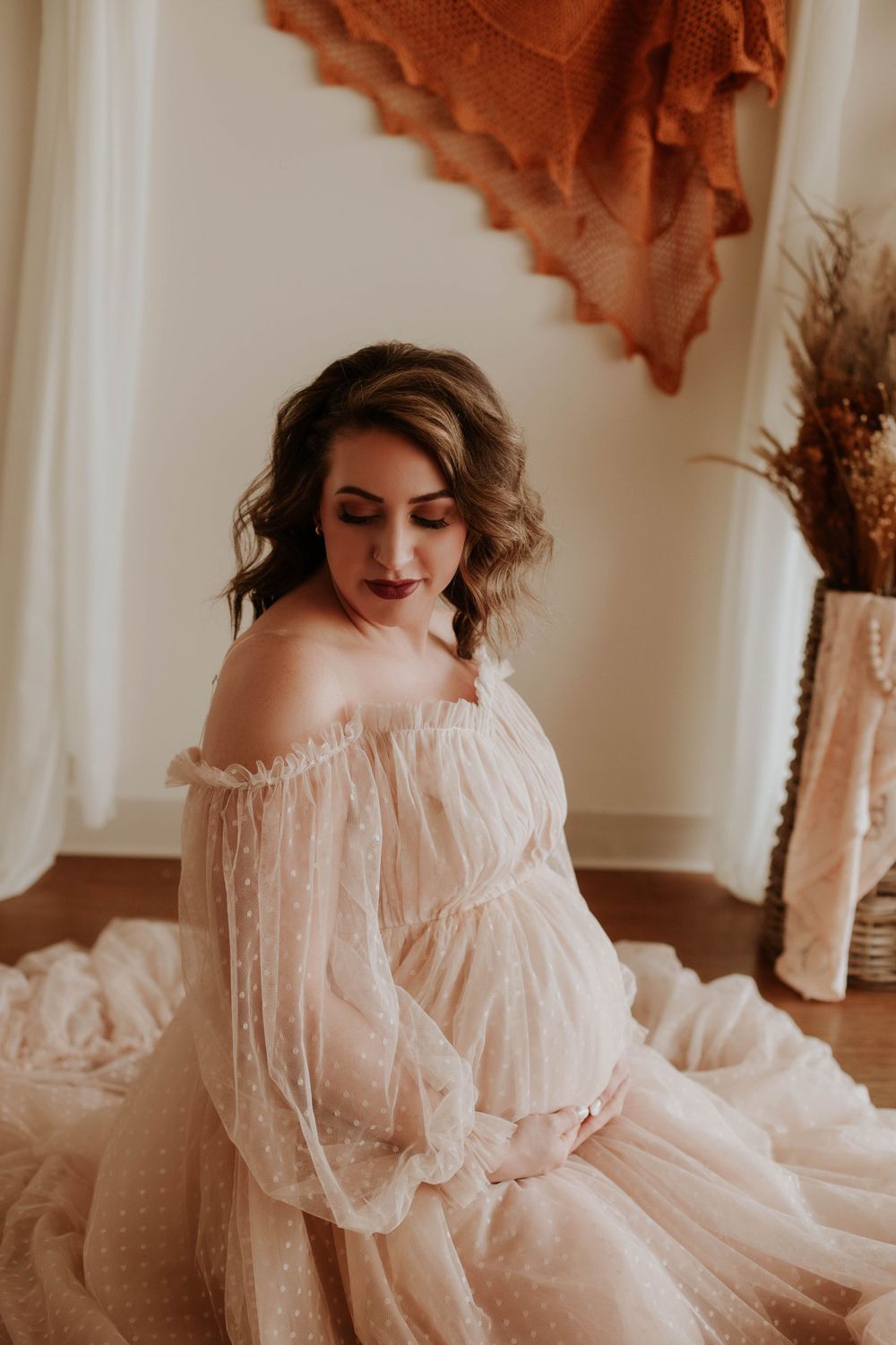 A pregnant woman in an off-shoulder blush dotted tulle gown sits gracefully in a boho studio.