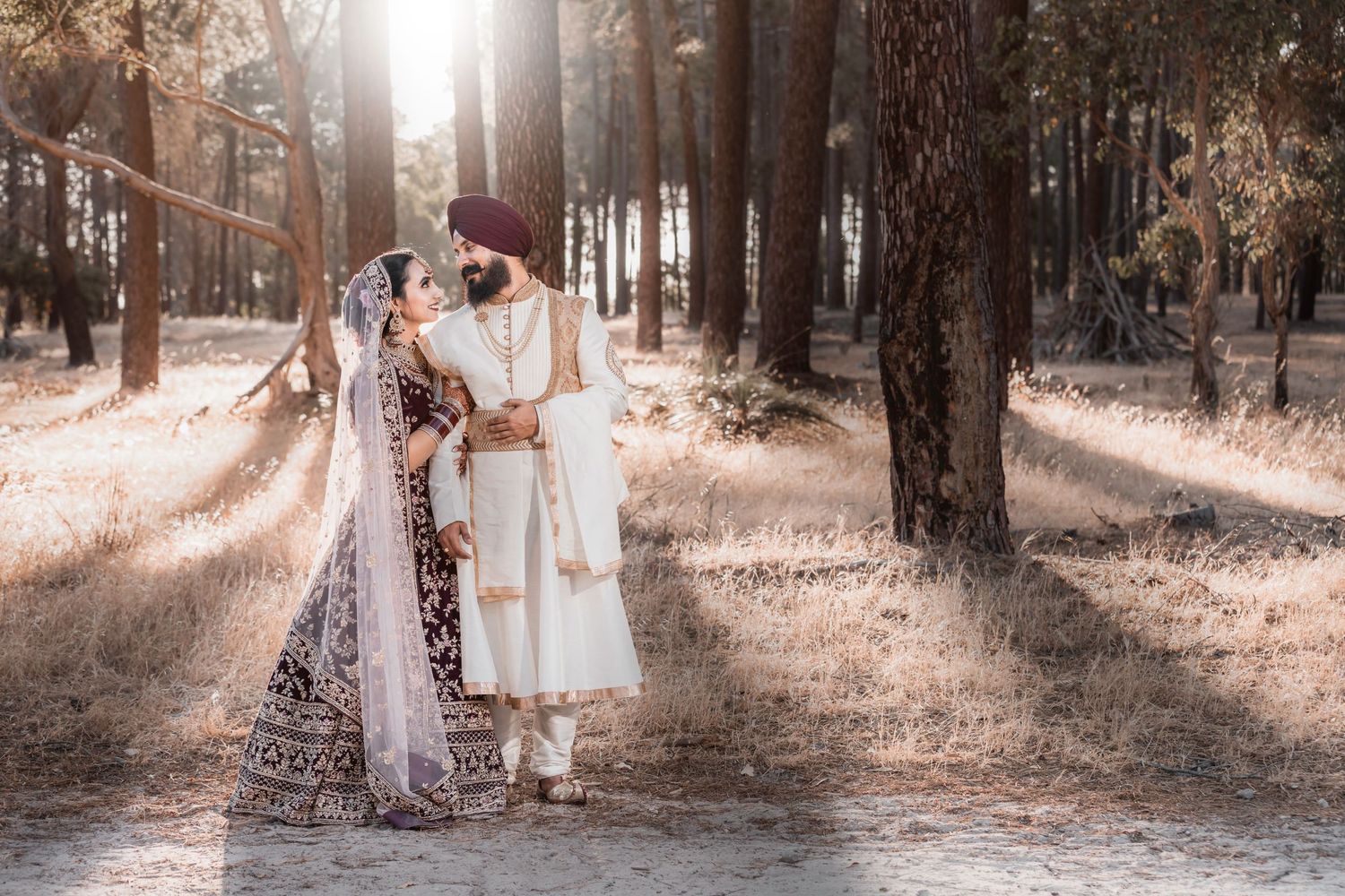 Romantic wedding couple embraces in sunlit forest wearing traditional and modern attire.