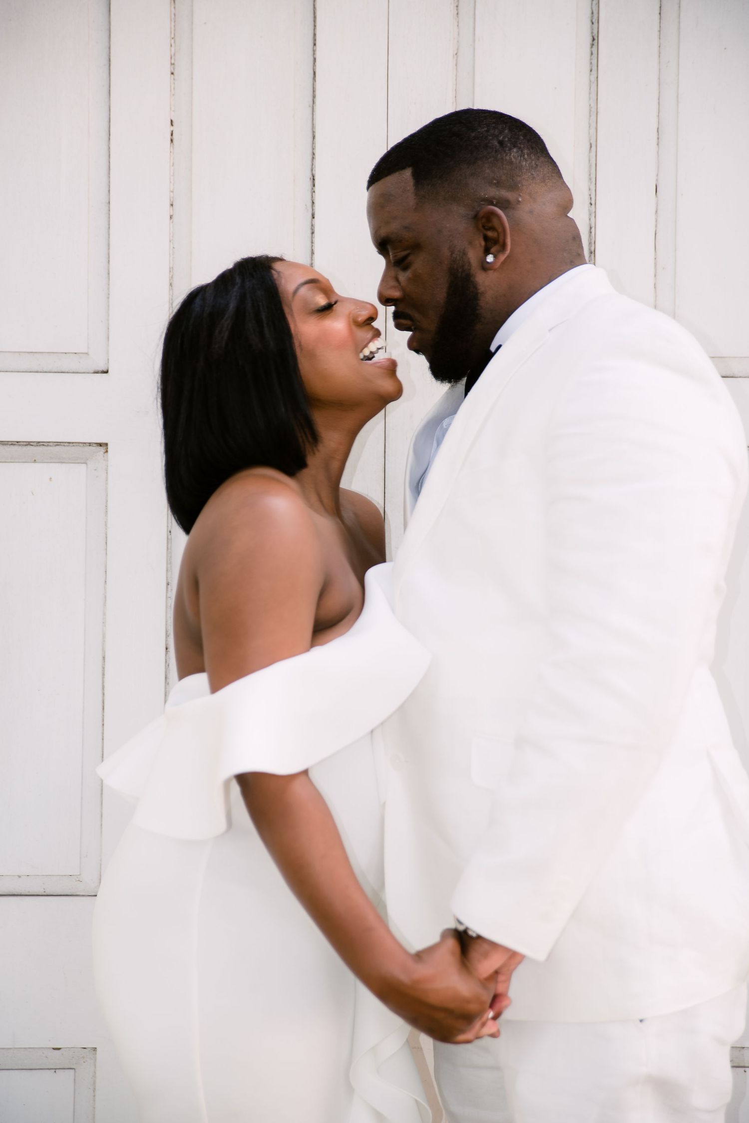 A romantic couple in white formal wear share intimate moments against a white wooden backdrop.