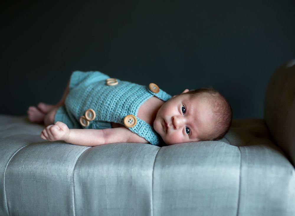 Baby in teal onesie lying on gray couch cushion.