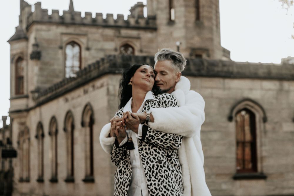 A couple in coordinating leopard print and white coats poses in front of a castle tower.