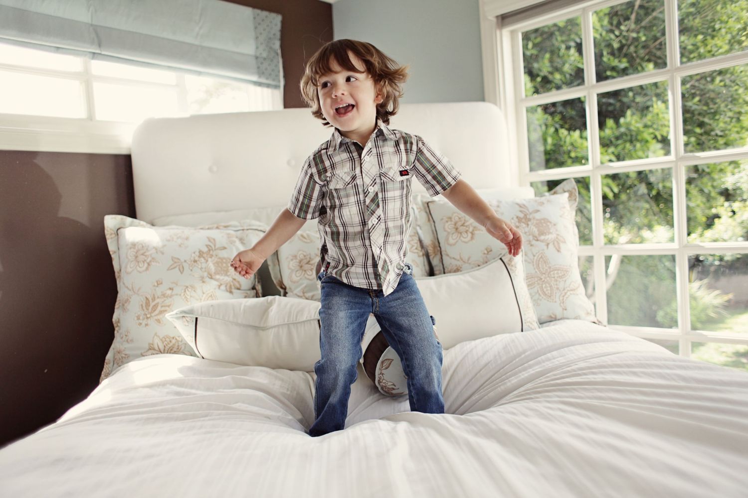 Young child jumping playfully on a white bed near a sunny window.